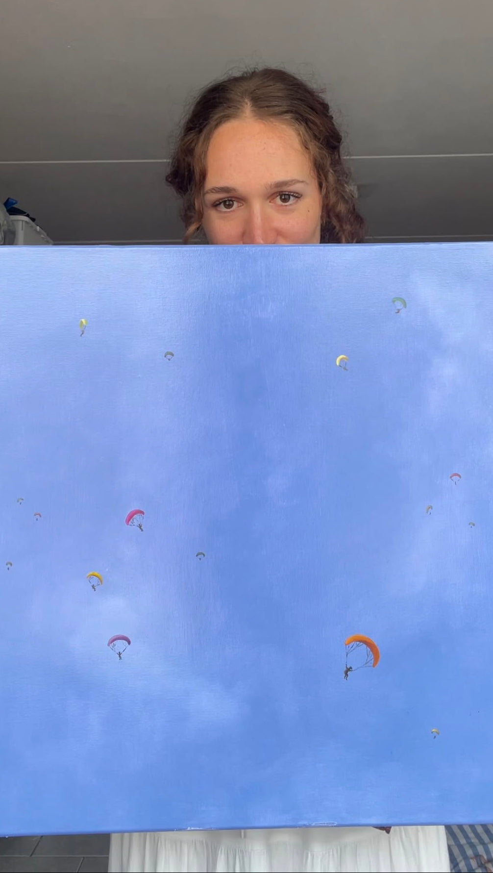 A person holding up a square canvas painting featuring a light blue sky with small colorful parachutes, with their face partially visible above the artwork.